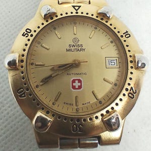 May include: A gold-toned Swiss Military automatic wristwatch. The watch features a gold dial with black numerals, a date window at the 3 o'clock position, and a red and white cross emblem. The watch has a gold bezel with raised markers.