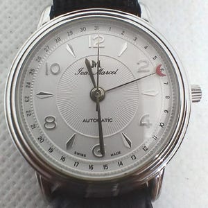 Może przedstawiać: Srebrny automatyczny zegarek na rękę Jean Marcel z czarnym skórzanym paskiem. Tarcza zegarka ma teksturowaną srebrną tarczę z czarnymi wskazówkami i cyframi. Zegarek wyświetla również datę i wskaźnik faz księżyca.