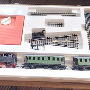 Puede incluir: Un juego de trenes modelo en una caja blanca. El juego incluye una locomotora negra y roja, dos vagones verdes, un panel de control rojo y varias piezas de vía. Para los entusiastas de los trenes modelo.