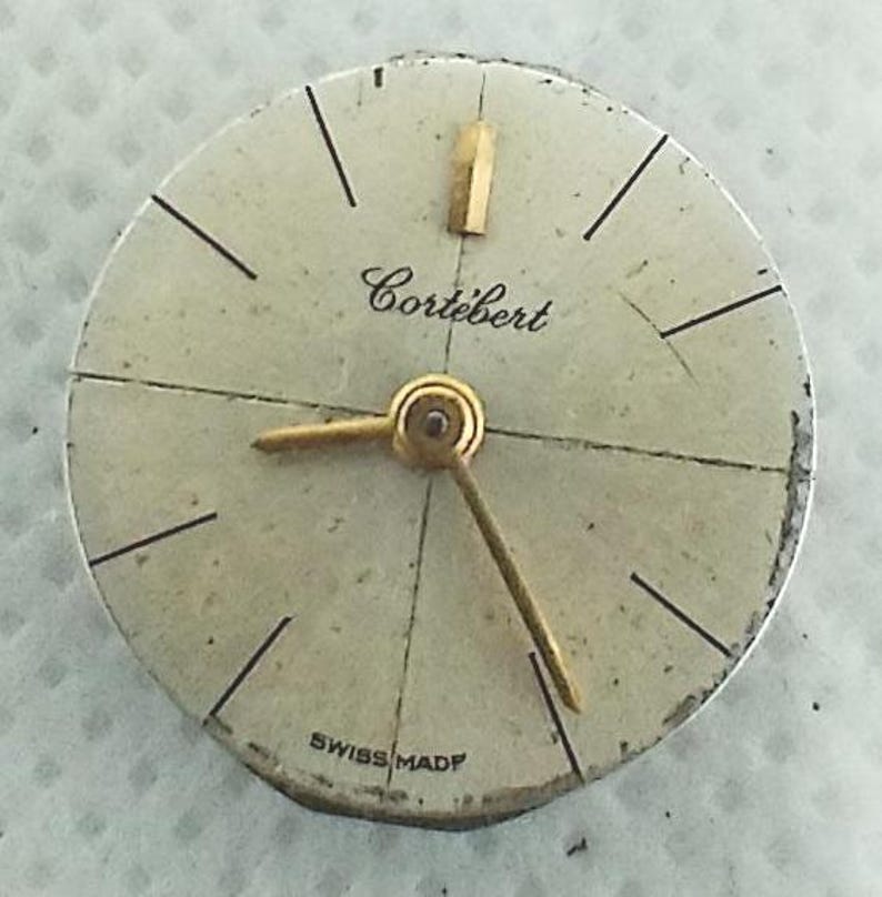 Op de afbeelding: Vintage horlogekast met de merknaam "Cortebert" in cursief schrift. De wijzerplaat is gebroken wit met zwarte uurmarkeringen en goudkleurige wijzers. De woorden "Swiss Made" staan onderaan.
