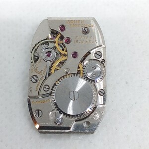 May include: A close-up of a Gruen Watch Co. watch movement. The silver-toned mechanism features gears, jewels, and the text "FIFTEEN 15 JEWELS". The watch movement is rectangular with rounded edges.