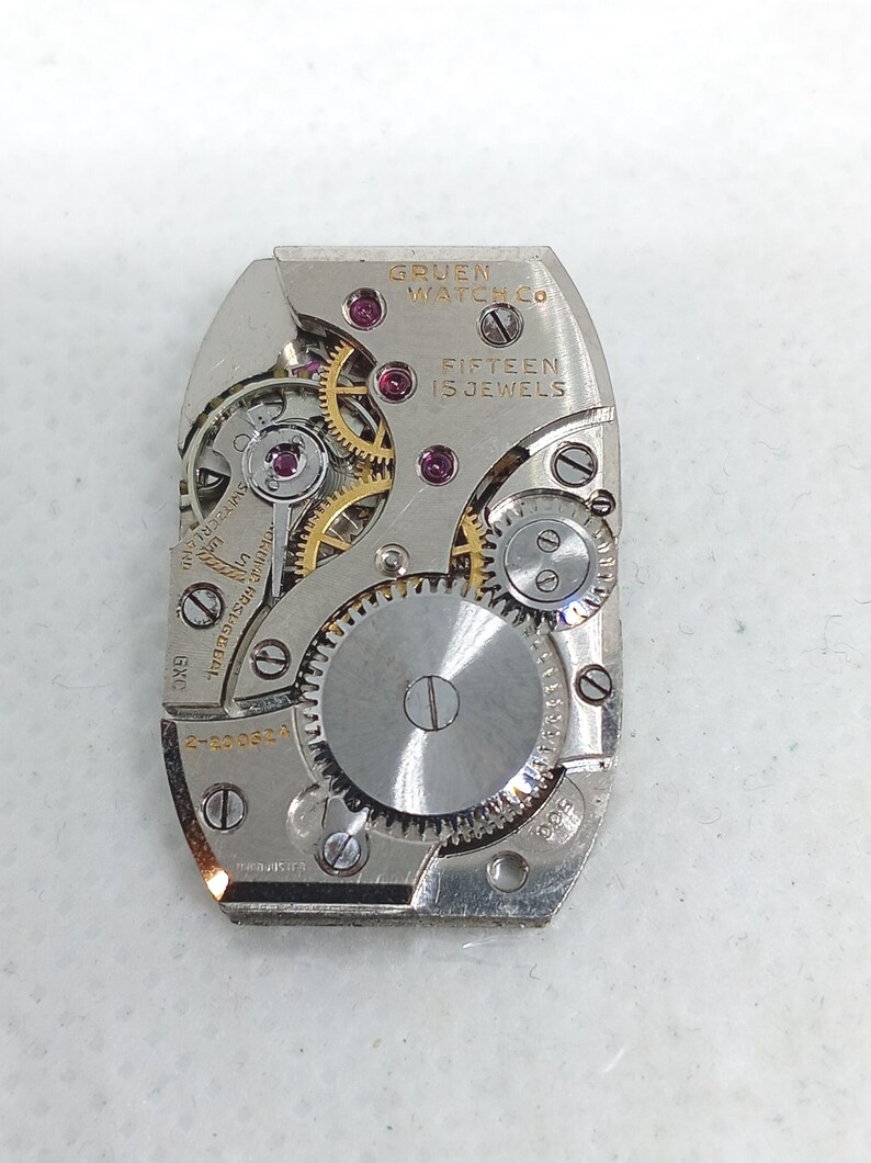 May include: A close-up of a Gruen Watch Co. watch movement. The silver-toned mechanism features gears, jewels, and the text "FIFTEEN 15 JEWELS". The watch movement is rectangular with rounded edges.