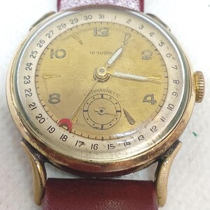 May include: A gold-tone wristwatch with a circular gold dial, featuring Arabic numerals at 2, 4, 8, 10, and 12. The outer rim displays a date calendar from 1 to 31. It includes a sub-dial, a red-tipped date hand, and a dark red leather strap.