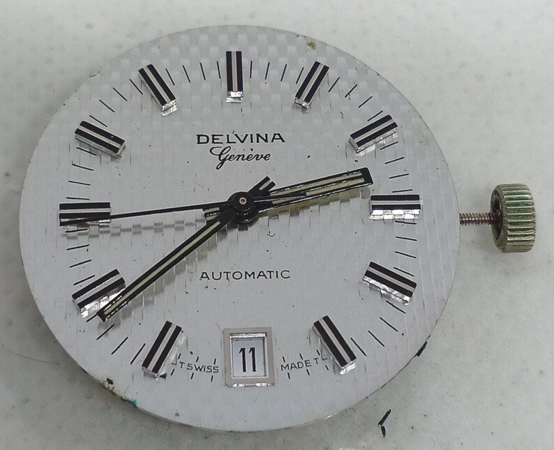 May include: A silver watch face with the text "DELVINA Geneve AUTOMATIC". The watch face features black and silver hour markers and hands. The date is displayed as "11". A green crown is visible on the right side.