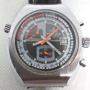 Könnte beinhalten: Eine silberfarbene Vintage-Armbanduhr mit schwarzem und grauem Zifferblatt. Die Uhr verfügt über einen orangefarbenen Sekundenzeiger, eine Datumsanzeige und mehrere Hilfszifferblätter. Das Zifferblatt trägt den Text "MEIS ANKER RUBIS 13".