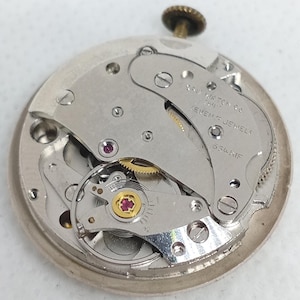 May include: A detailed view of a vintage watch movement, showcasing its complex internal workings. The silver mechanism includes ruby jewels and the text "SWISS WATCH CO SEVEN JEWELS 534 KIF". A small, gold crown is at the top.