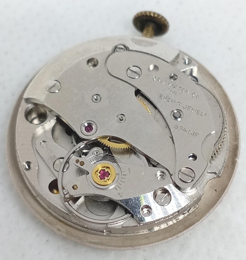May include: A detailed view of a vintage watch movement, showcasing its complex internal workings. The silver mechanism includes ruby jewels and the text "SWISS WATCH CO SEVEN JEWELS 534 KIF". A small, gold crown is at the top.