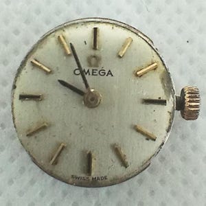 Op de afbeelding: Een vintage Omega horlogekast met een cr&egrave;mekleurige wijzerplaat, goudkleurige wijzers en markeringen en een gouden kroon. Het woord "OMEGA" staat bovenaan gedrukt en "SWISS MADE" onderaan. De wijzerplaat is rond.