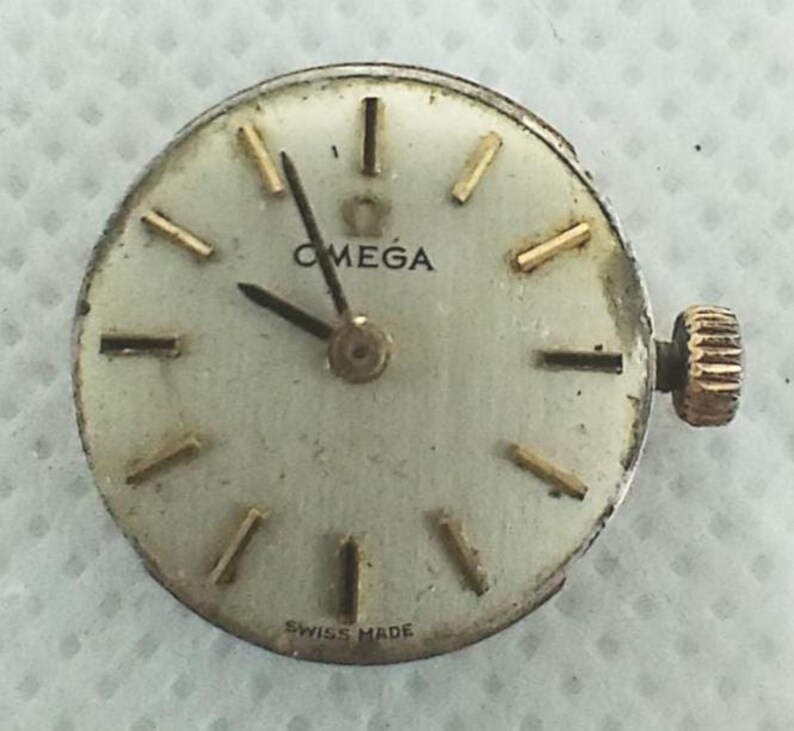 Op de afbeelding: Een vintage Omega horlogekast met een cr&egrave;mekleurige wijzerplaat, goudkleurige wijzers en markeringen en een gouden kroon. Het woord "OMEGA" staat bovenaan gedrukt en "SWISS MADE" onderaan. De wijzerplaat is rond.