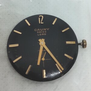 May include: A black watch face with gold-colored hands and markers. The text "CAUNY SWISS LORD" is printed in gold. The watch face has a gold-colored crown on the right side.