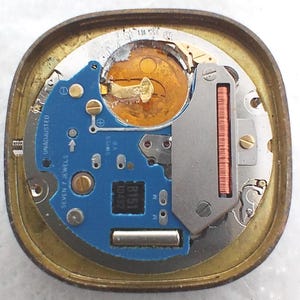 May include: A close-up view of a watch movement, showcasing intricate internal components. The movement features a blue circuit board with the text "SEVEN 7 JEWELS" and "UNADJUSTED". A gold-colored battery and various metal parts are visible within the gold-toned case.