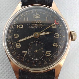 May include: A vintage gold-coloured Anker wristwatch with a black face and a black leather strap. The watch face displays Arabic numerals, a subdial, and the text "ANKER 21 RUBIS STOSSGESICHERT WASSERDICHT" and "ANTIMAGNETIC". It has a date indicator.
