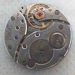 May include: Close-up of a Piaget watch movement. The circular mechanism features various gears, jewels, and a balance wheel. The watch movement is silver-toned with gold-colored accents and the Piaget logo is visible.