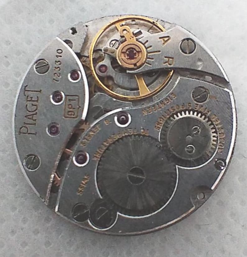 May include: Close-up of a Piaget watch movement. The circular mechanism features various gears, jewels, and a balance wheel. The watch movement is silver-toned with gold-colored accents and the Piaget logo is visible.