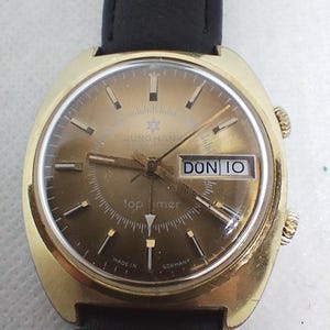 May include: A gold-toned Junghans Top Timer watch with a black leather band. The watch face is a warm brown color with gold markers and hands. The day and date are displayed in a window. The watch is marked "Made in Germany."