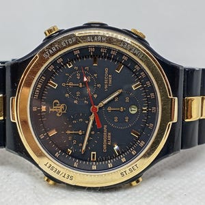 Op de afbeelding: Zwart en goudkleurig polshorloge met een donkerblauwe wijzerplaat. Het horloge beschikt over chronograaf-, alarm- en timerfuncties. De horlogeband is zwart met gouden accenten. De wijzerplaat toont de tekst "TACHYMETER", "CHRONOGRAPH ALARM" en "1/50 SECOND TIMER".