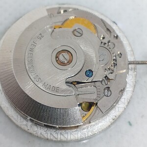May include: Detailed view of a silver watch movement, showcasing complex gears and parts. The mechanism is labelled "25 JEWELS SWISS MADE". The design incorporates gold and blue details, with a screw attached to the side.