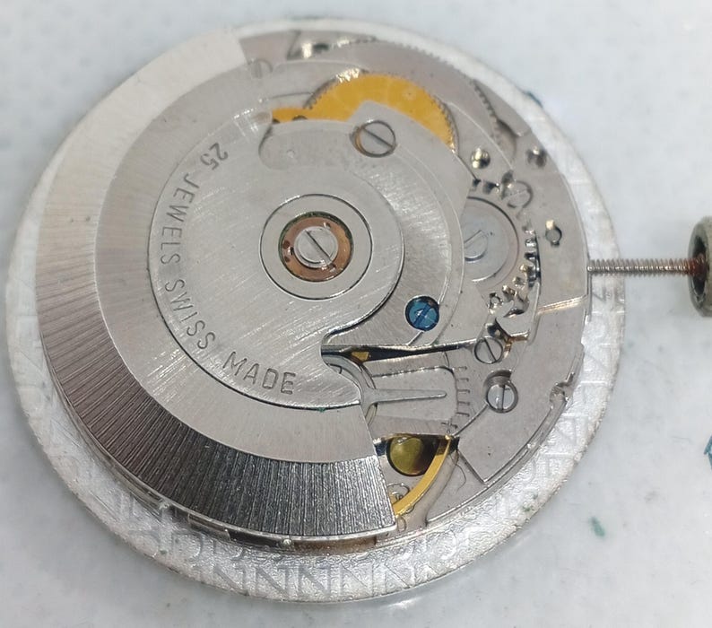May include: Detailed view of a silver watch movement, showcasing complex gears and parts. The mechanism is labelled "25 JEWELS SWISS MADE". The design incorporates gold and blue details, with a screw attached to the side.