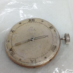 May include: A vintage Omega Automatic watch face with Roman numeral hour markers and hands. The watch face is silver with the Omega logo and "Automatic" text. A small, textured crown is visible on the side.