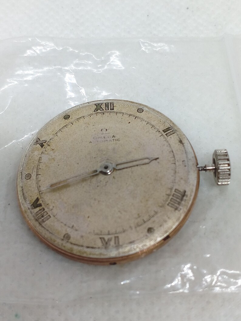 May include: A vintage Omega Automatic watch face with Roman numeral hour markers and hands. The watch face is silver with the Omega logo and "Automatic" text. A small, textured crown is visible on the side.