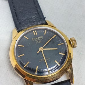 Może przedstawiać: Zegarek Phenix w stylu vintage w kolorze złotym z czarną tarczą i czarnym skórzanym paskiem. Tarcza zegarka posiada złote wskazówki i znaczniki z napisem "PHENIX 17 JEWELS Stop". Zegarek posiada również złotą koronkę.