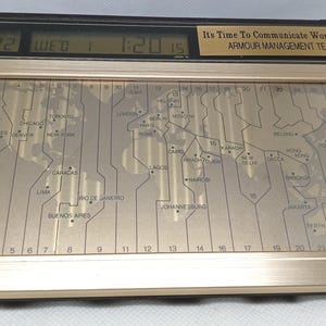 May include: A gold-coloured world time clock with a digital display showing the date and time. The clock face features a world map with time zones and city names. The text "Its Time To Communicate Worldwide. ARMOUR MANAGEMENT TEAM" is displayed.