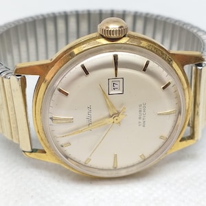 Peut inclure: Une montre vintage dorée avec un cadran argenté et un affichage de la date à 3 heures. La montre présente des aiguilles et des index dorés, et le cadran est marqué "Alina" et "17 Rubis Antichoc". Le bracelet est extensible et argenté.