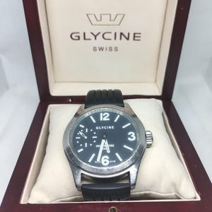 Puede incluir: Un reloj suizo Glycine con esfera negra, bisel plateado y correa de caucho negro. El reloj se muestra en una caja de madera. La esfera tiene números y agujas blancas, con las palabras "Glycine" y "Swiss" visibles.
