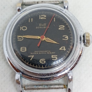 Könnte beinhalten: Vintage Armbanduhr mit schwarzem Zifferblatt, silberfarbenem Gehäuse und goldfarbener Krone. Die Uhr hat goldfarbene Zeiger und Ziffern, mit einem roten Sekundenzeiger. Auf dem Zifferblatt steht "EWZ 20 RUBIS" und "ANTIMAGNETIC SUPER SHOCK RESIST".