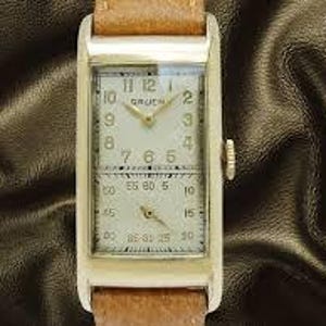 May include: A rectangular gold-toned Gruen wristwatch with a light-colored face and a brown leather band. The watch face has two sections, one for the time and the other for seconds. The watch has a crown on the right side.