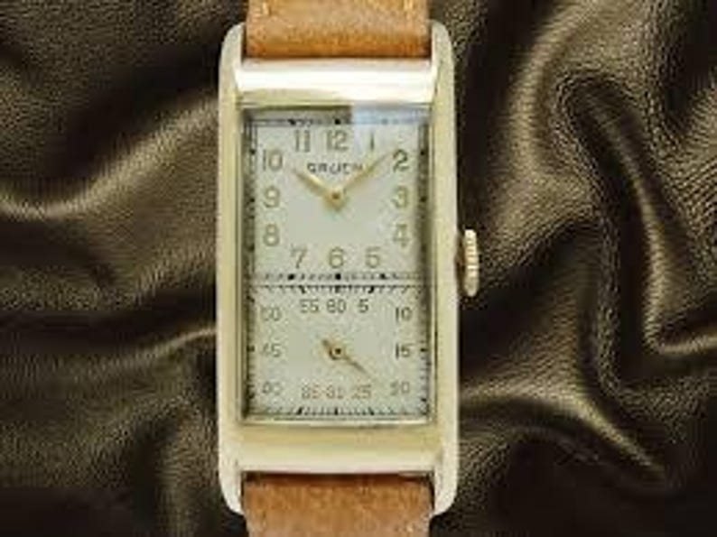 May include: A rectangular gold-toned Gruen wristwatch with a light-colored face and a brown leather band. The watch face has two sections, one for the time and the other for seconds. The watch has a crown on the right side.