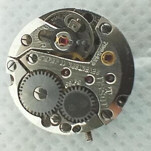 Op de afbeelding: Close-up van een uurwerk met ingewikkelde tandwielen, schroeven en juwelen. Het zilverkleurige mechanisme toont de merknaam "Tissot" en de tekst "Seventen 17 Jewels Swiss". Het ronde uurwerk is gedetailleerd.