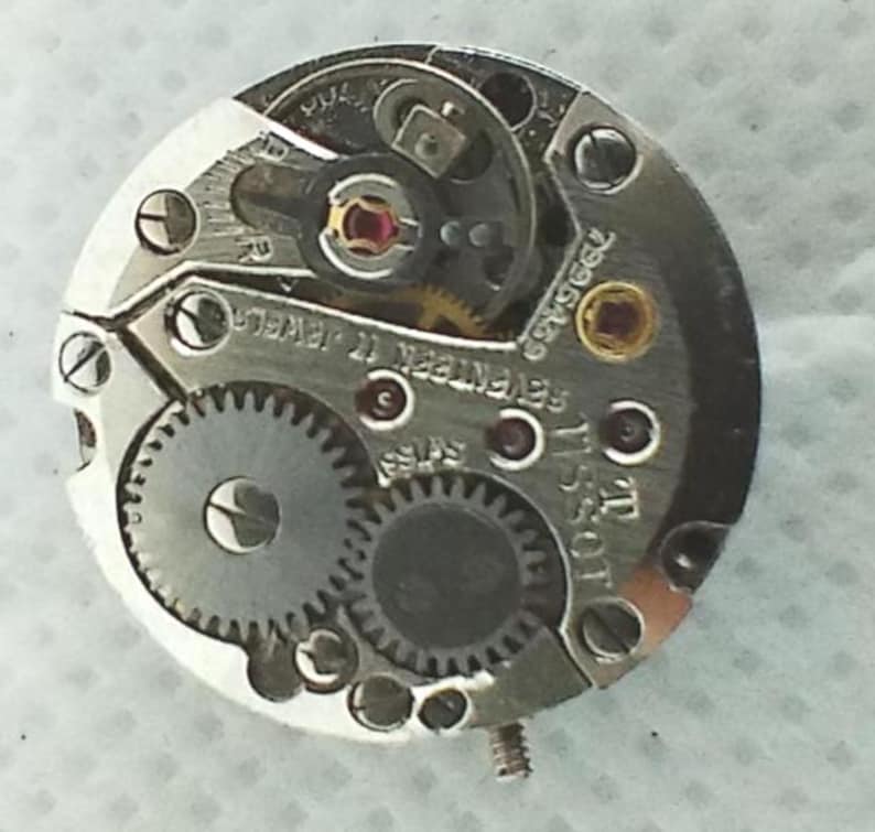Op de afbeelding: Close-up van een uurwerk met ingewikkelde tandwielen, schroeven en juwelen. Het zilverkleurige mechanisme toont de merknaam "Tissot" en de tekst "Seventen 17 Jewels Swiss". Het ronde uurwerk is gedetailleerd.