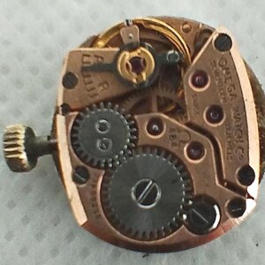 Op de afbeelding: Close-up van een Omega Watch Co. uurwerk. Het mechanisme toont goudkleurige metalen componenten, waaronder tandwielen, schroeven en een opwindas. De tekst "OMEGA WATCH CO." en "17 JEWELS" zijn zichtbaar. Een complex voorbeeld van horologische techniek.