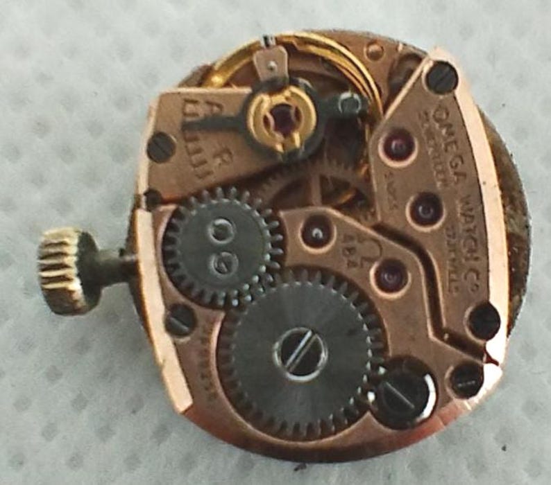 Op de afbeelding: Close-up van een Omega Watch Co. uurwerk. Het mechanisme toont goudkleurige metalen componenten, waaronder tandwielen, schroeven en een opwindas. De tekst "OMEGA WATCH CO." en "17 JEWELS" zijn zichtbaar. Een complex voorbeeld van horologische techniek.