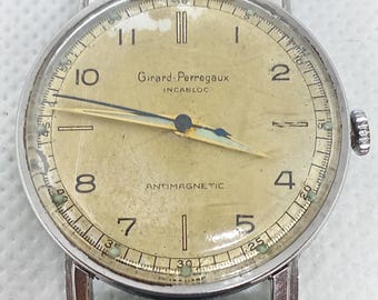 Könnte beinhalten: Eine Vintage-Armbanduhr von Girard-Perregaux mit silberfarbenem Gehäuse und schwarzem Lederarmband. Das Zifferblatt der Uhr ist hellgoldfarben mit schwarzen Ziffern und Zeigern. Der Text "Girard-Perregaux" und "ANTIMAGNETIC" ist sichtbar.