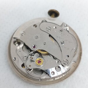 May include: Detailed view of a vintage watch mechanism, revealing the internal workings. The silver-coloured gears, jewels, and plates are displayed, with a small crown at the top. The text "SEVENTEEN JEWELS" is visible on the movement.