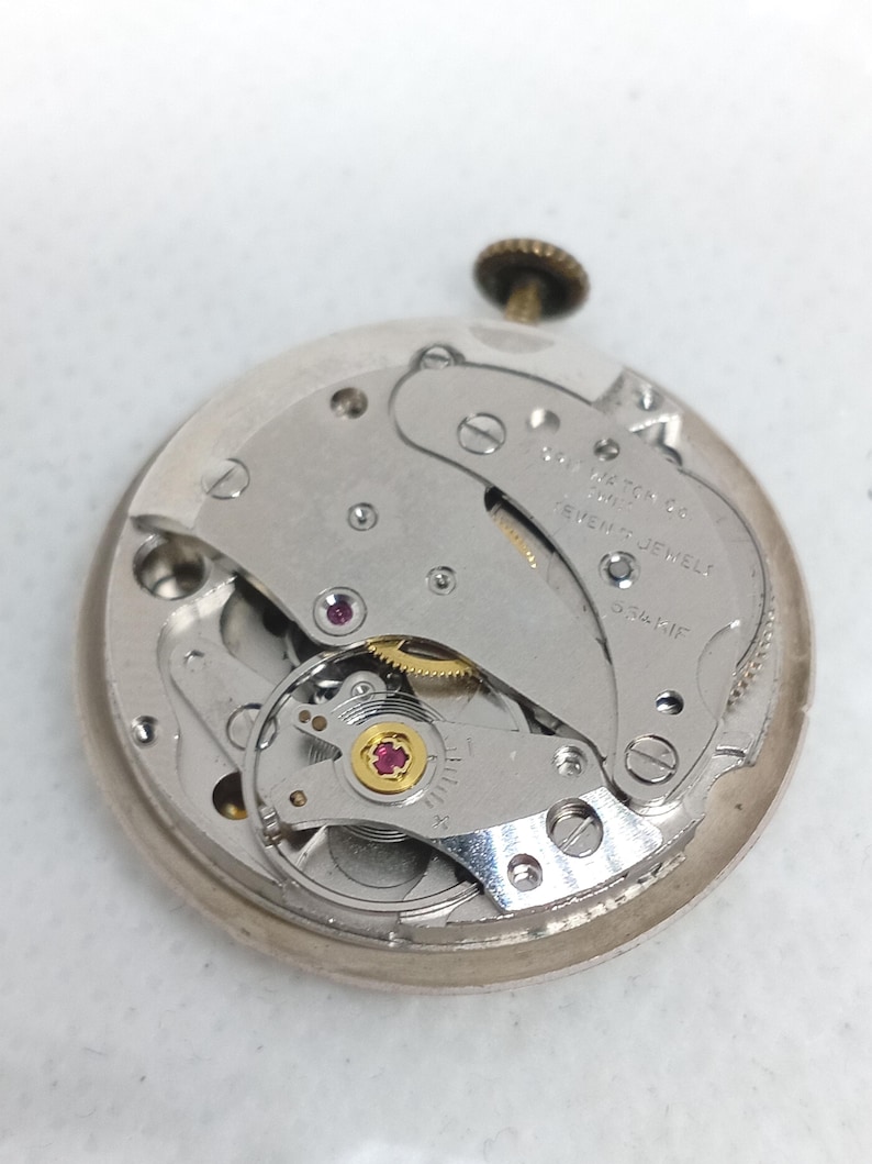 May include: Detailed view of a vintage watch mechanism, revealing the internal workings. The silver-coloured gears, jewels, and plates are displayed, with a small crown at the top. The text "SEVENTEEN JEWELS" is visible on the movement.