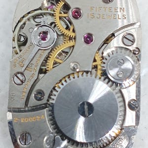May include: Close-up of a Gruen Watch Co. watch movement. The silver-toned mechanism features gold gears, ruby jewels, and the text "FIFTEEN 15 JEWELS". The watch part has several screws and the number "2-200634".