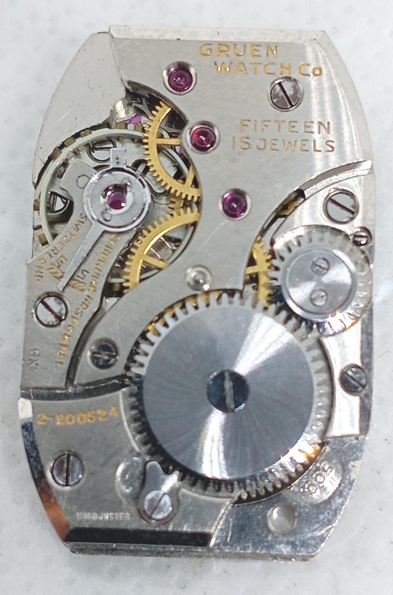 May include: Close-up of a Gruen Watch Co. watch movement. The silver-toned mechanism features gold gears, ruby jewels, and the text "FIFTEEN 15 JEWELS". The watch part has several screws and the number "2-200634".