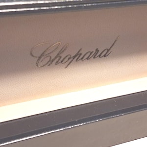 May include: A beige interior lining of a box featuring the brand name Chopard embossed in a cursive script. The box has a dark, smooth exterior border at the bottom and a matching dark edge at the top, creating a clean and structured appearance.