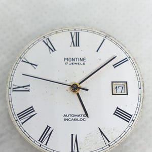 Może przedstawiać: Biała tarcza zegarka z czarnymi rzymskimi cyframi i wskaz&oacute;wkami. Tarcza zegarka zawiera napis "MONTINE 17 JEWELS AUTOMATIC INCABLOC" oraz okienko daty z numerem 17. Tarcza zegarka jest okrągła.