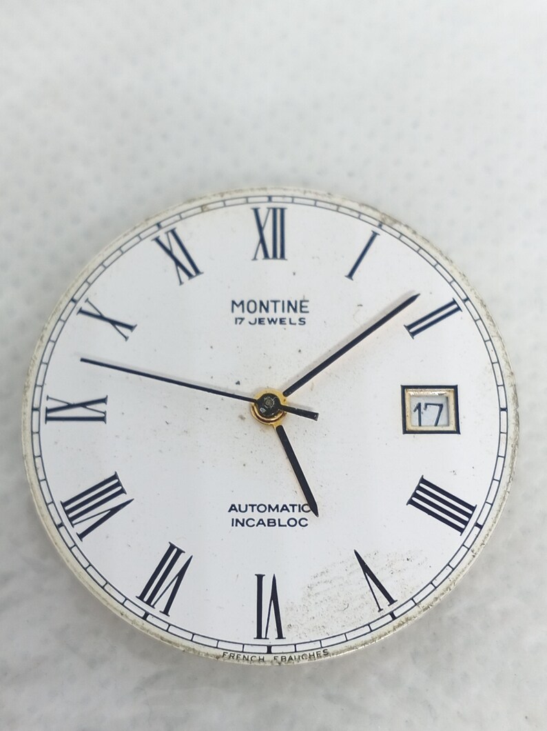 Może przedstawiać: Biała tarcza zegarka z czarnymi rzymskimi cyframi i wskaz&oacute;wkami. Tarcza zegarka zawiera napis "MONTINE 17 JEWELS AUTOMATIC INCABLOC" oraz okienko daty z numerem 17. Tarcza zegarka jest okrągła.
