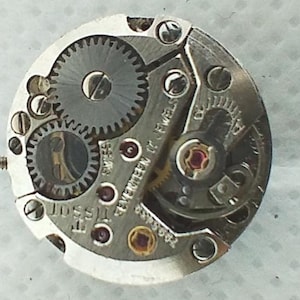 Op de afbeelding: Close-up van een Tissot uurwerk. Het zilverkleurige mechanisme heeft meerdere tandwielen, juwelen en schroeven. De tekst "Tissot" en "Seventeen 17 Jewels" zijn zichtbaar. Het uurwerk is een complex stuk horologische techniek.