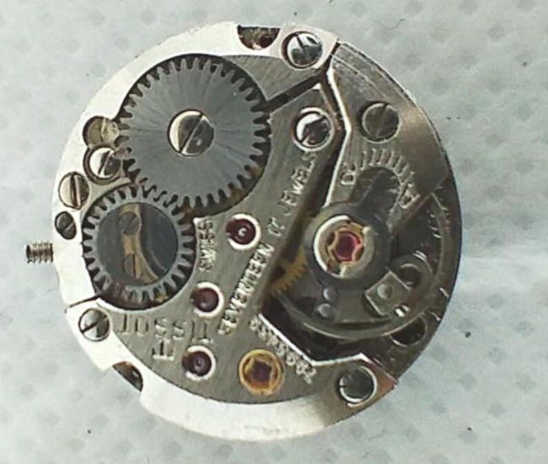 Op de afbeelding: Close-up van een Tissot uurwerk. Het zilverkleurige mechanisme heeft meerdere tandwielen, juwelen en schroeven. De tekst "Tissot" en "Seventeen 17 Jewels" zijn zichtbaar. Het uurwerk is een complex stuk horologische techniek.