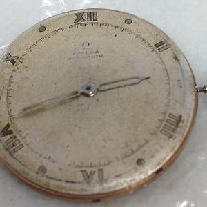 May include: A vintage Omega Automatic watch face with Roman numeral hour markers and a small seconds hand. The watch face is a light, aged color with a silver-tone metal casing and a winding crown on the side. The watch face has the Omega logo.