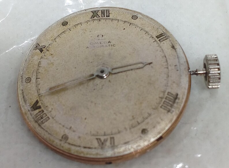May include: A vintage Omega Automatic watch face with Roman numeral hour markers and a small seconds hand. The watch face is a light, aged color with a silver-tone metal casing and a winding crown on the side. The watch face has the Omega logo.