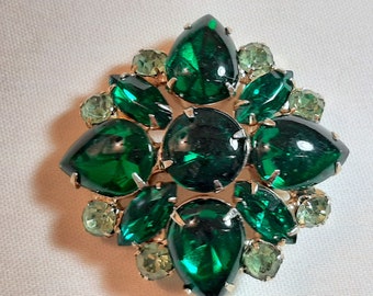 Pode incluir: Broche vintage com uma moldura dourada, apresentando um grupo central de grandes strass verde-esmeralda, em forma de gota e redondos. Strass menores, verde claro, circundam o grupo central, criando um design floral ou em estrela. O broche está sobre um fundo branco.