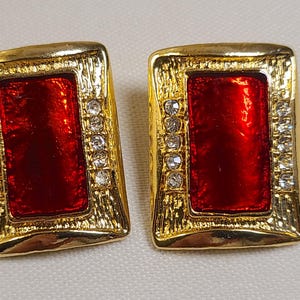 May include: A pair of rectangular earrings featuring a central glossy red panel framed by a textured gold-tone border. Each earring is accented with a vertical row of five clear, round-cut rhinestones along both the left and right sides.