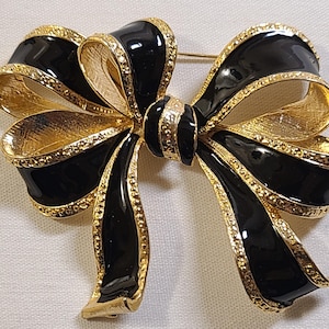 May include: A gold and black bow brooch with a textured gold finish and black enamel. The bow is tied in a knot with the ends of the bow pointing out to the sides.